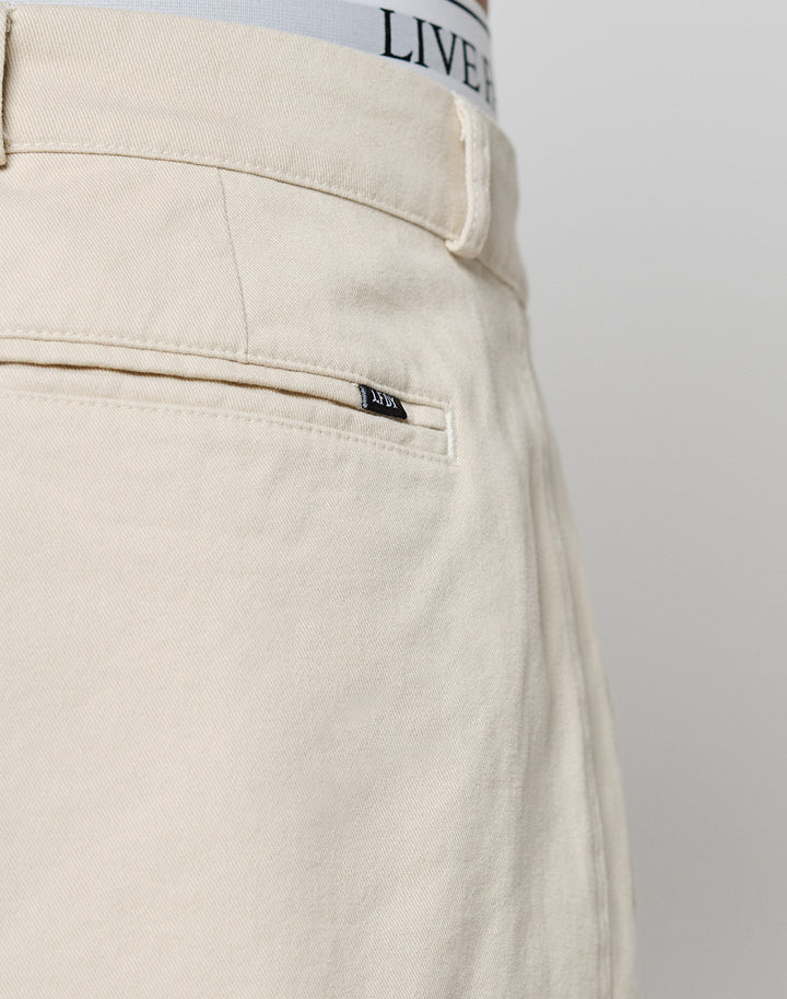 Close-up of the back of beige Wide Chino pants by LIVE FAST DIE YOUNG, in 100% cotton, showing a welt pocket with a black tag, part of a belt loop, and the waistband of LIVE FAST DIE YOUNG underwear visible at the top.