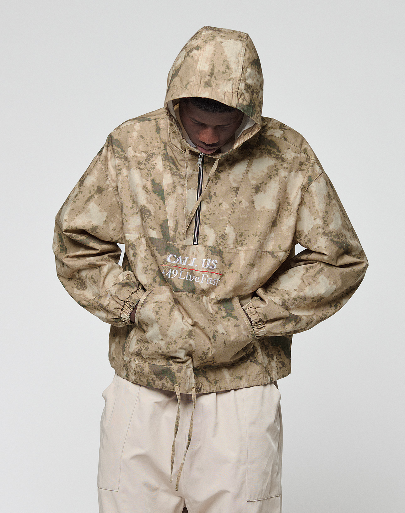 A person wears the LFDY Camo Halfzip Jacket by LIVE FAST DIE YOUNG, a tan and beige camo hooded jacket with "CALL US 49 Live Fast" on the front and an oversize fit. Hands in pockets, they face down against a plain white background.