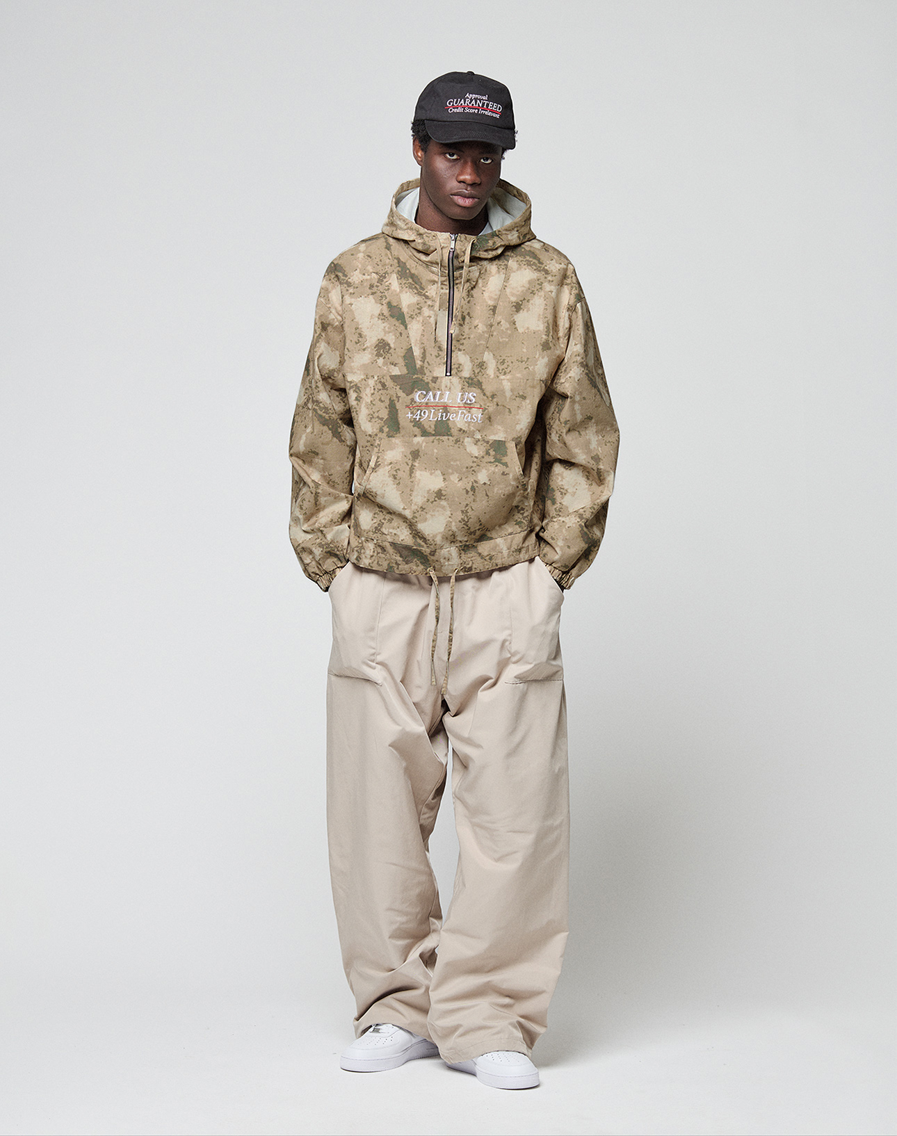 A person wears LIVE FAST DIE YOUNG Baggy Nylon Pants, a tan camo hoodie, white sneakers, and a dark cap, standing against a plain light background with hands in pockets, looking at the camera.