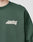 A person wears the LIVE FAST DIE YOUNG Basic Chest Sweater, a dark green oversize fit made from a soft cotton-polyester blend, featuring the brand’s bold white logo on the left chest. Only their upper torso and neck are shown.