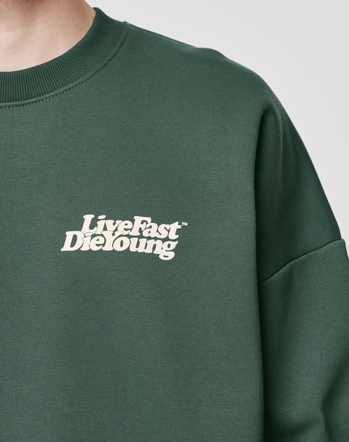 A person wears the LIVE FAST DIE YOUNG Basic Chest Sweater, a dark green oversize fit made from a soft cotton-polyester blend, featuring the brand’s bold white logo on the left chest. Only their upper torso and neck are shown.
