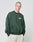 A person with a shaved head wears the LIVE FAST DIE YOUNG Basic Chest Sweater, an oversize fit dark green sweatshirt with white chest text, and light beige shorts. They stand against a white background, hands in pockets, looking at the camera.