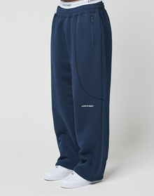 Person wearing LFDY Active Fleece Jogger by LIVE FAST DIE YOUNG—wide fit navy sweatpants made from soft Baumwolle with side zip pocket, curved seams, LIVE FAST logo on thigh, white waistband, and white sneakers—standing against a light background.