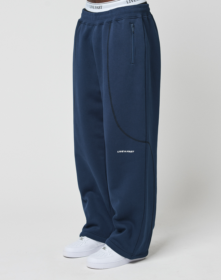 Person wearing LFDY Active Fleece Jogger by LIVE FAST DIE YOUNG—wide fit navy sweatpants made from soft Baumwolle with side zip pocket, curved seams, LIVE FAST logo on thigh, white waistband, and white sneakers—standing against a light background.