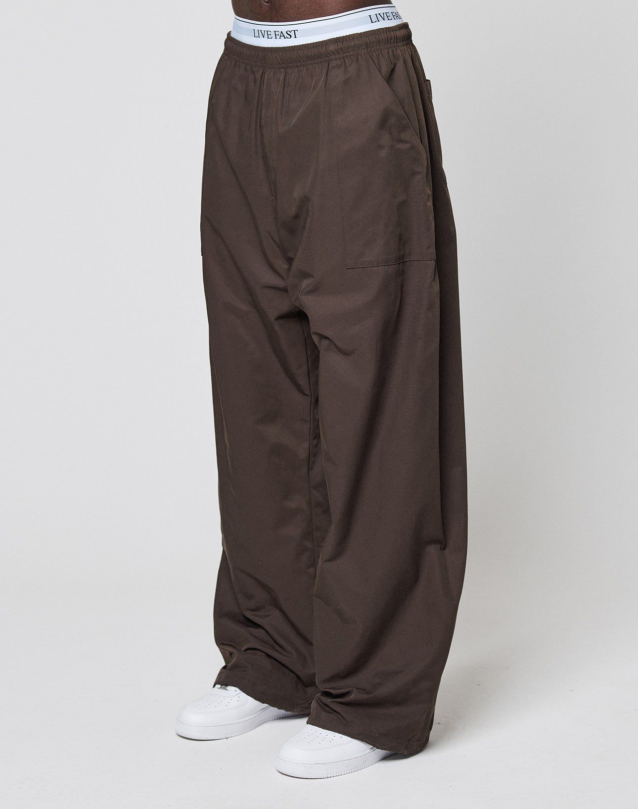 A person wearing LIVE FAST DIE YOUNG Baggy Nylon Pants with an elastic waistband and white sneakers stands against a plain white background, with the "LIVE FAST" text visible on the waistband.
