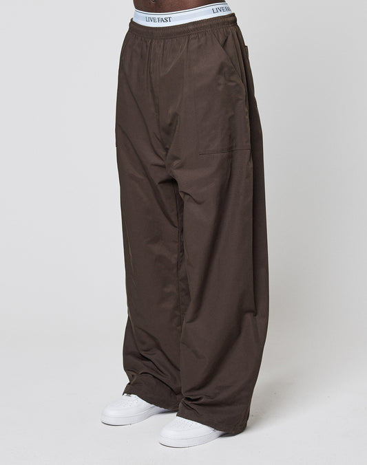 A person wearing LIVE FAST DIE YOUNG Baggy Nylon Pants with an elastic waistband and white sneakers stands against a plain white background, with the "LIVE FAST" text visible on the waistband.