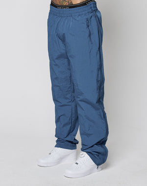A person wearing blue LIVE FAST DIE YOUNG Zip Tape Track Pants with an elastic waistband, white sneakers, and black underwear with a visible LIVE FAST DIE YOUNG waistband stands against a plain white background.