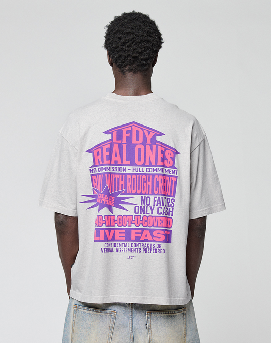 A person with short curly hair faces away, wearing the LIVE FAST DIE YOUNG LFDY Real Ones Tee—an oversize 100% Baumwolle T-shirt in light gray, featuring bold purple and pink graphic slogans like “LFDY REAL ONES” and a subtle flaglabel on the back.