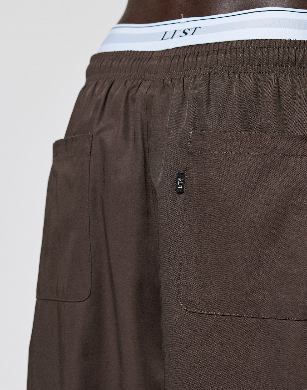 Close-up of a person wearing LIVE FAST DIE YOUNG Baggy Nylon Pants with large back pockets, showing the white LIST-labeled underwear waistband peeking out at the waist.