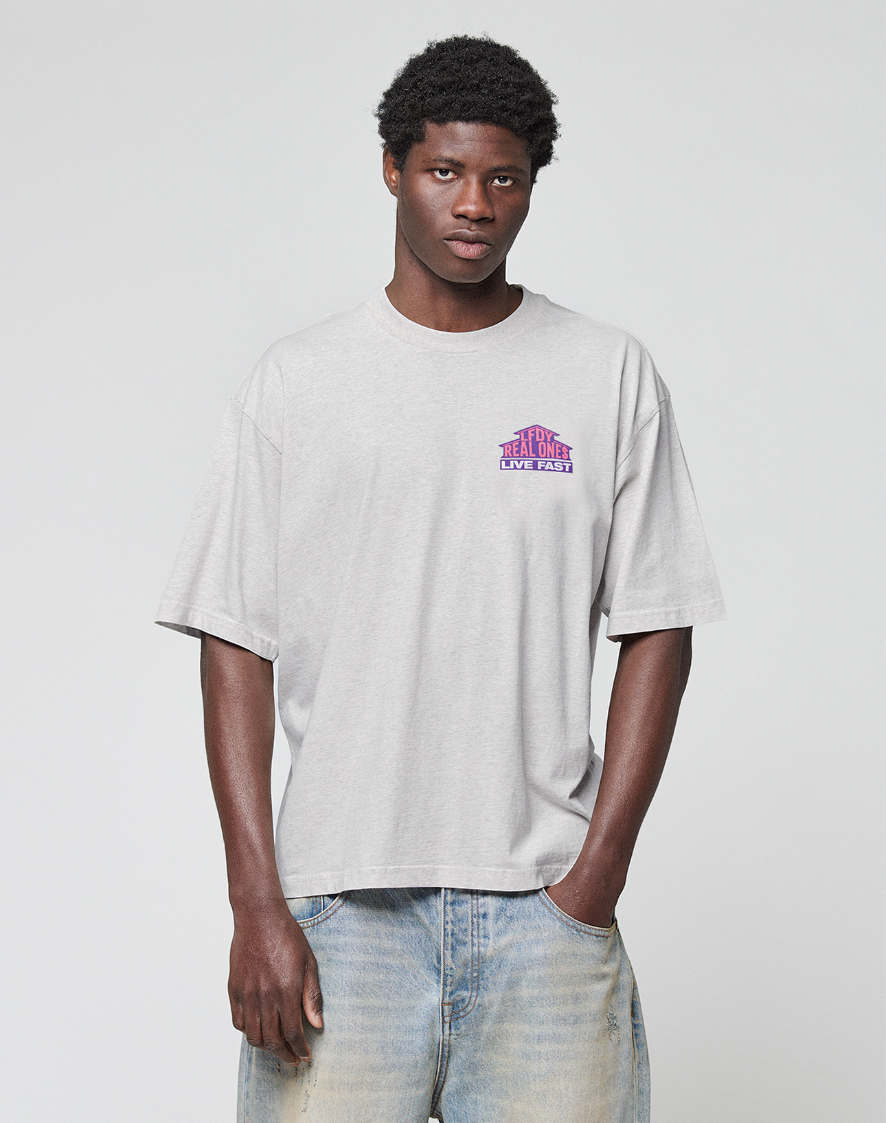 A person wears the LIVE FAST DIE YOUNG LFDY Real Ones Tee—light gray with pink and purple text—styled in an oversize fit with faded blue jeans, one hand in their pocket, posing against a plain white background.