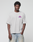 A person wears the LIVE FAST DIE YOUNG LFDY Real Ones Tee—light gray with pink and purple text—styled in an oversize fit with faded blue jeans, one hand in their pocket, posing against a plain white background.