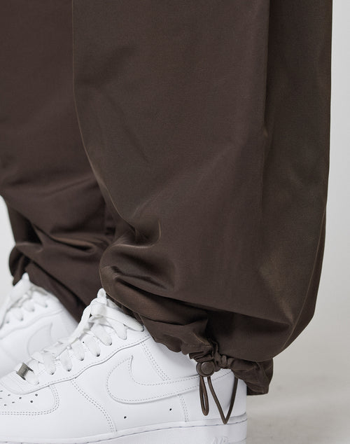 Close-up of a person wearing LIVE FAST DIE YOUNG Baggy Nylon Pants in brown, featuring a wide fit, elastic waistband, ankle drawstrings, zip pockets, and gathered cuffs. The pants are styled with white Nike sneakers.