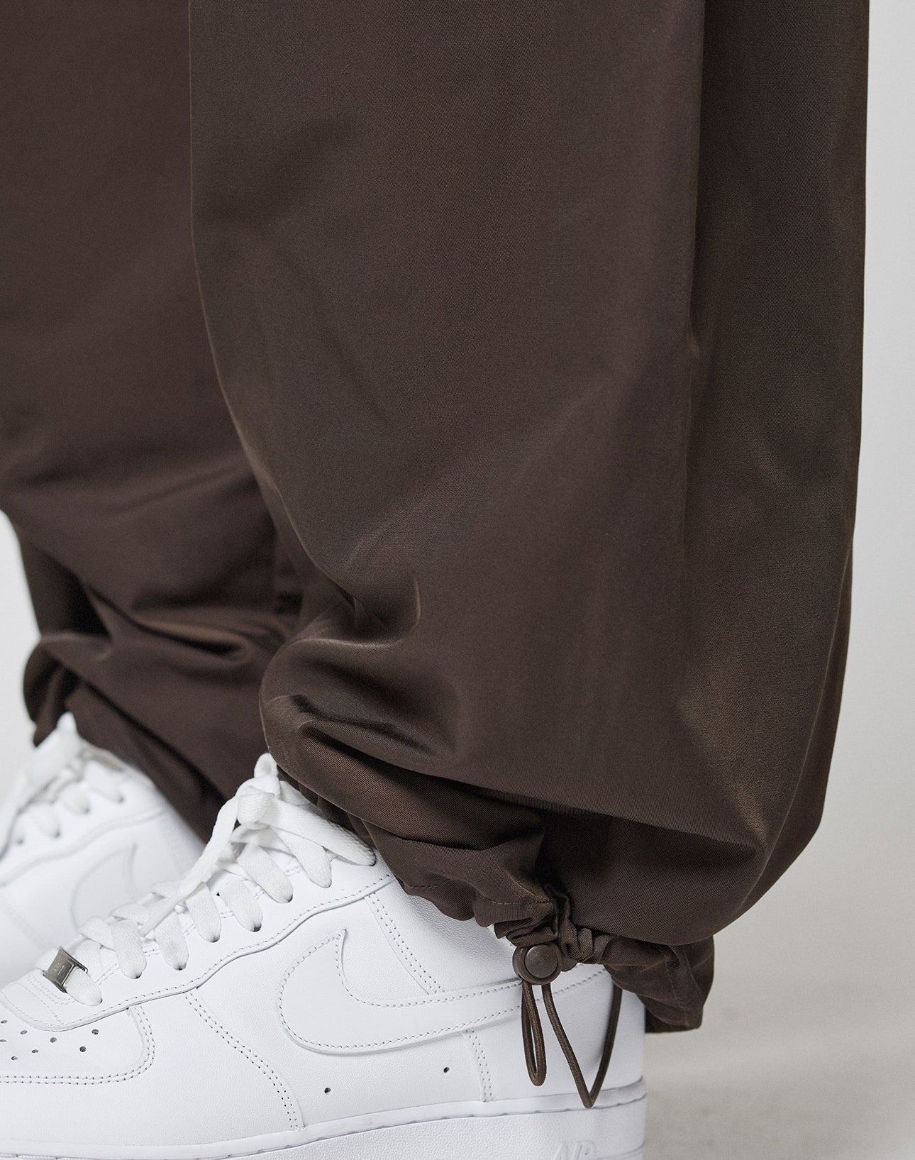 Close-up of a person wearing LIVE FAST DIE YOUNG Baggy Nylon Pants in brown, featuring a wide fit, elastic waistband, ankle drawstrings, zip pockets, and gathered cuffs. The pants are styled with white Nike sneakers.