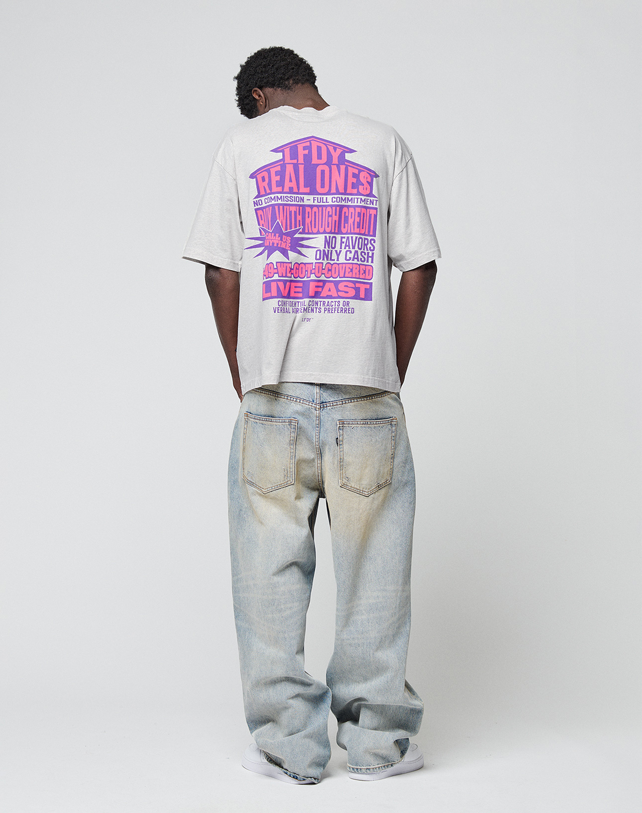 A person faces away wearing an oversized light gray LIVE FAST DIE YOUNG LFDY Real Ones Tee with bold pink and purple text, paired with baggy, faded blue jeans. Their hands are in their pockets against a plain white background.