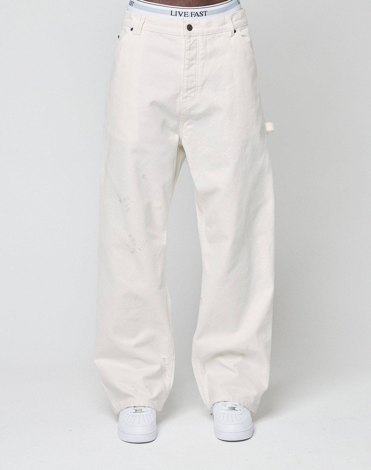 A model wears LIVE FAST DIE YOUNG Worker Pants—loose-fitting, off-white with a baggy fit, carpenter pocket, and branded white waistband—paired with white sneakers against a plain light background.