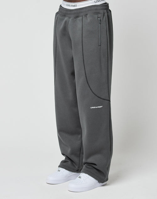 A person wears the LFDY Active Fleece Jogger by LIVE FAST DIE YOUNG in dark gray with zip pockets, a waistband featuring LIVE FAST text, and a small LIVE FAST logo on one leg, paired with white sneakers.