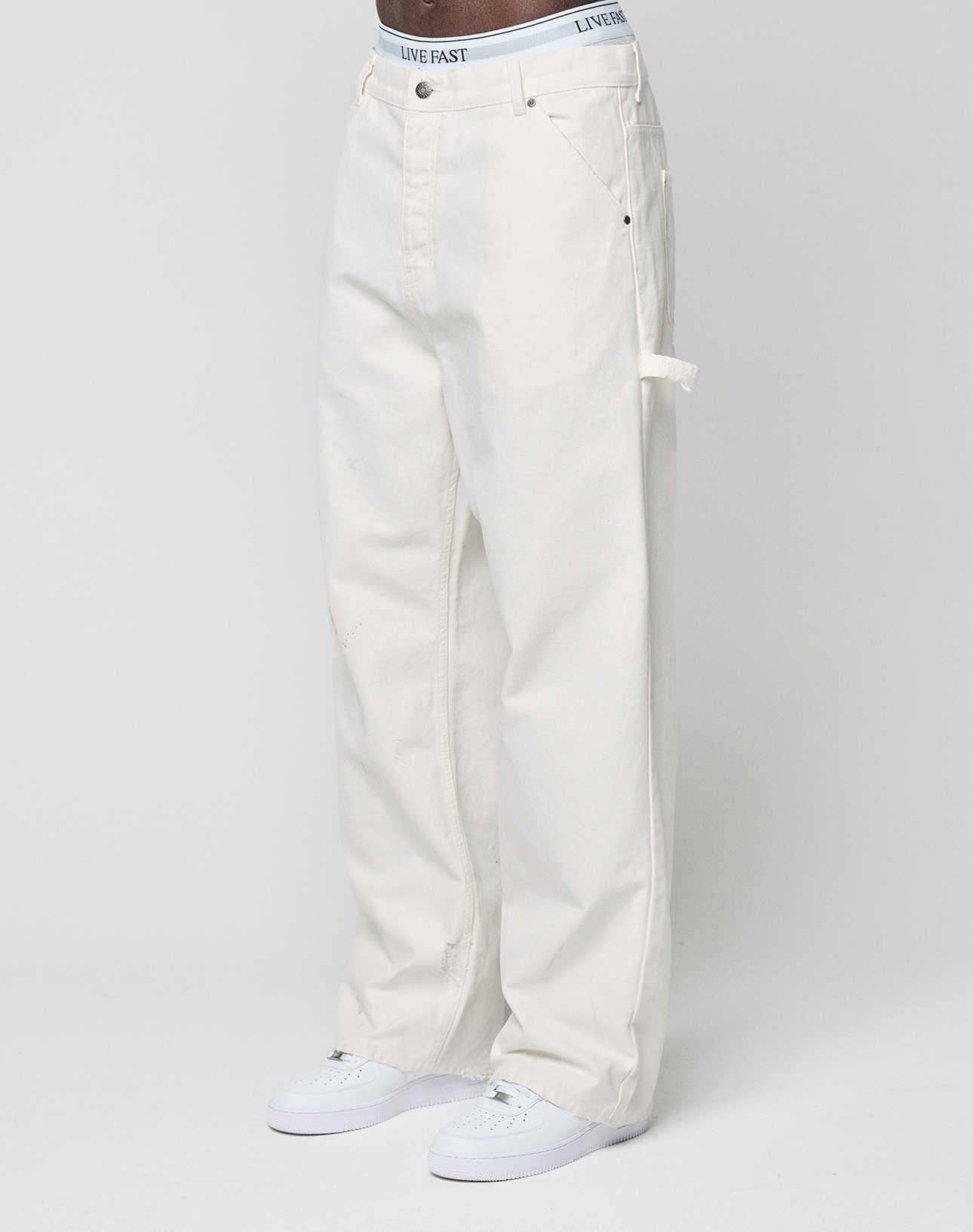 A person models LIVE FAST DIE YOUNG Worker Pants—loose-fit, off-white carpenter style with a carpenter pocket—paired with white sneakers and a waistband featuring LIVE FAST, standing against a plain light background.