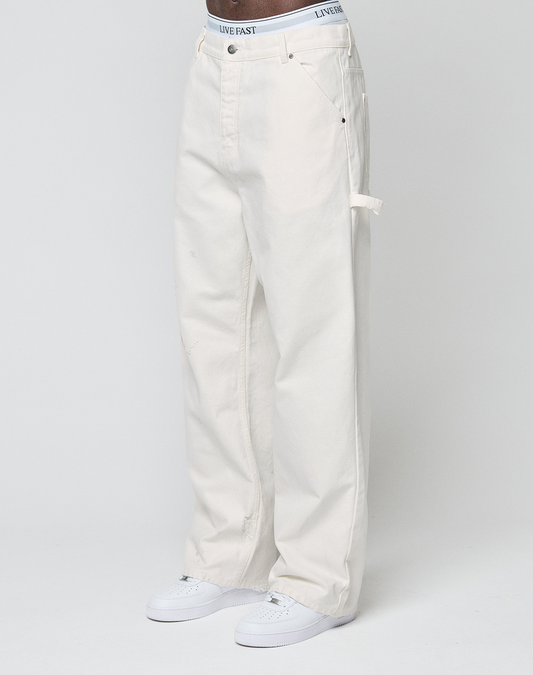 A person models LIVE FAST DIE YOUNG Worker Pants—loose-fit, off-white carpenter style with a carpenter pocket—paired with white sneakers and a waistband featuring LIVE FAST, standing against a plain light background.