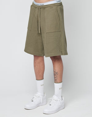 A person wears LIVE FAST DIE YOUNG Basic Knit Shorts in olive green with an elastic waistband, white LIVE FAST waistband underwear, white socks featuring LIVE FAST text, and white Nike sneakers against a plain light background.