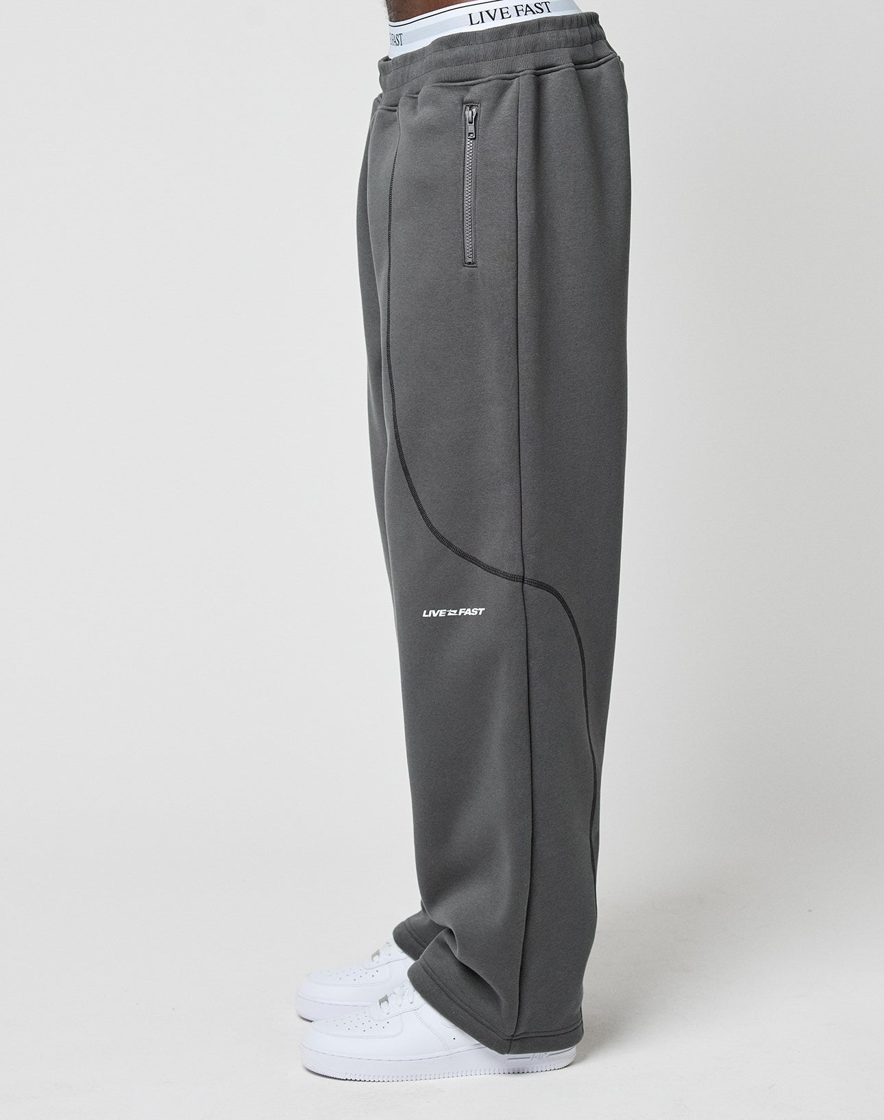 A person in LIVE FAST DIE YOUNG’s LFDY Active Fleece Jogger—dark gray sweatpants with a zip pocket and white LIVE FAST logo—paired with white sneakers, stands sideways against a plain white background.
