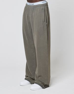 A person wears LFDY Ripped Sweatpants by LIVE FAST DIE YOUNG in olive green with a wide fit and elastic waistband, paired with white sneakers and LIVE FAST waistband underwear, standing against a plain light background.