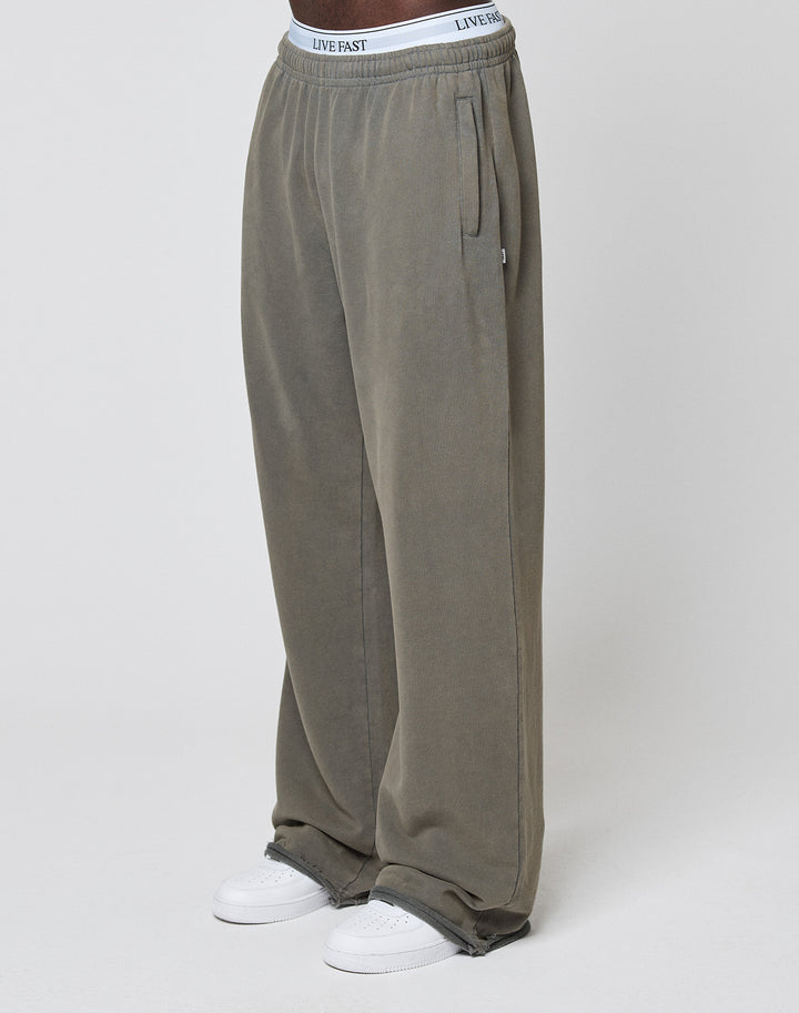 A person wears LFDY Ripped Sweatpants by LIVE FAST DIE YOUNG in olive green with a wide fit and elastic waistband, paired with white sneakers and LIVE FAST waistband underwear, standing against a plain light background. | model