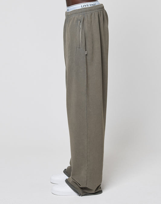 A person in LFDY Ripped Sweatpants by LIVE FAST DIE YOUNG—wide fit, olive green, 100% cotton, zipper pocket, white LIVE FAST waistband—with white sneakers, standing sideways against a plain white background.