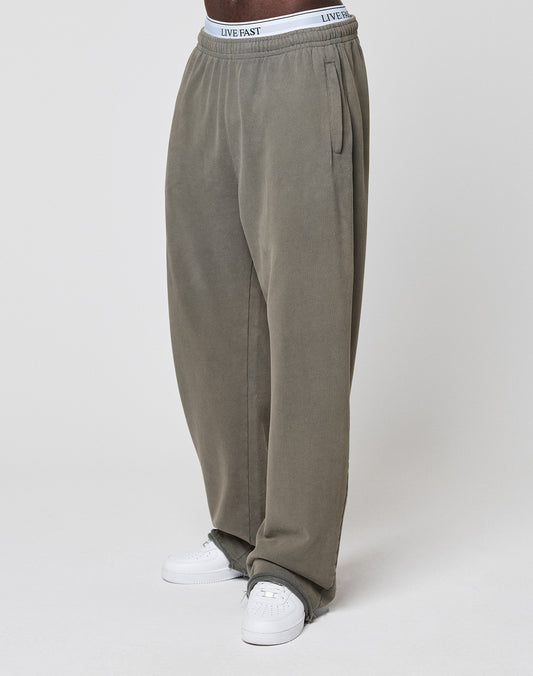 A person is standing against a plain white background, wearing LFDY Ripped Sweatpants from LIVE FAST DIE YOUNG in olive green wide fit with an elastic waistband, paired with white sneakers.