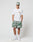 A man stands against a plain background wearing the LIVE FAST DIE YOUNG LFDY Thunder Chest Tee in white, paired with green and white tie-dye shorts, a matching cap, white crew socks, and sneakers. He looks straight ahead with hands in his pockets.