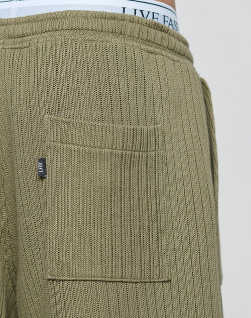 Close-up of olive green Basic Knit Shorts by LIVE FAST DIE YOUNG, featuring a ribbed texture, elastic waistband, and a rectangular back pocket with a black tag. The waistband of white underwear with LIVE FAS text is visible above the shorts.