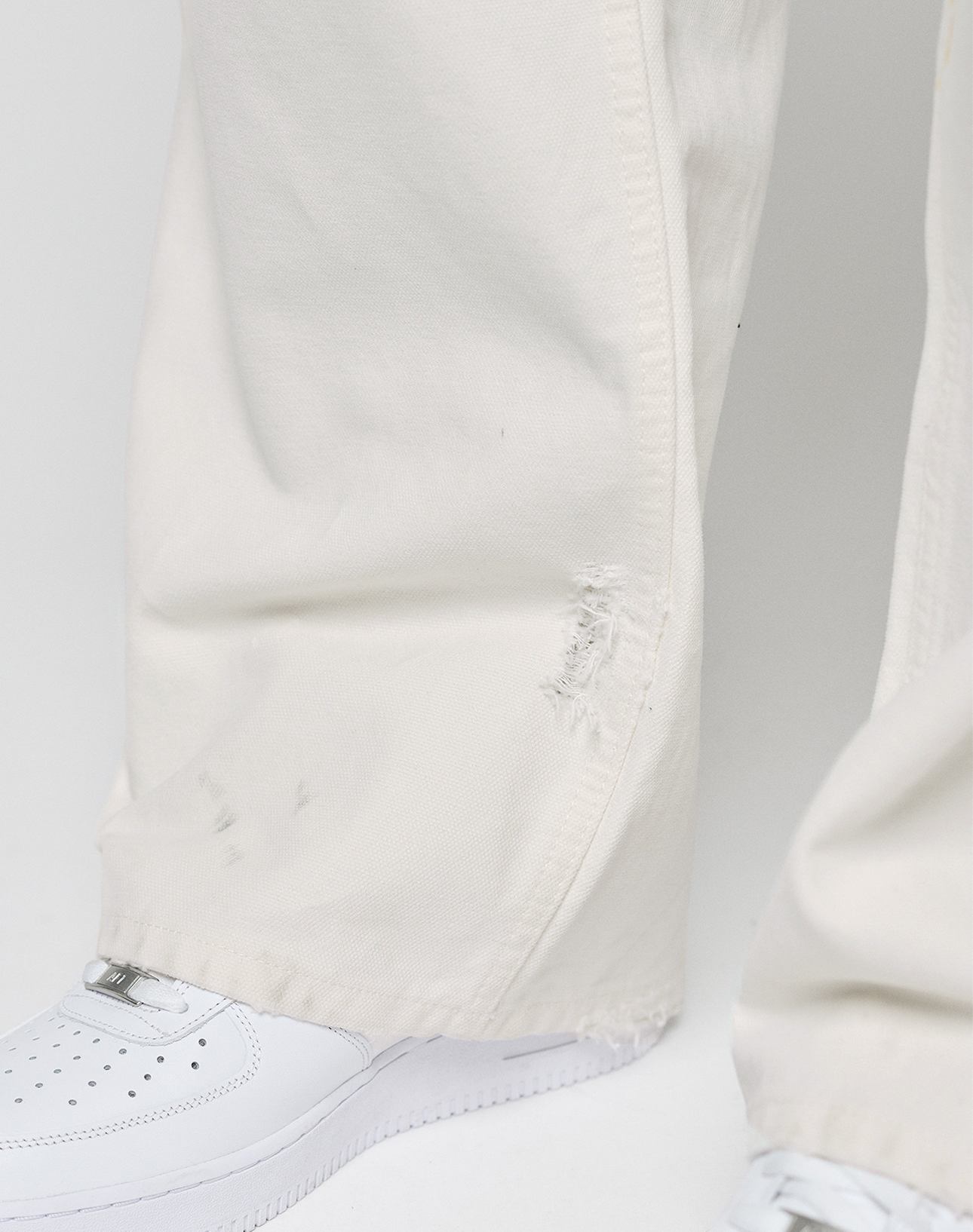 Close-up of a person wearing LIVE FAST DIE YOUNG Worker Pants in off-white with a baggy fit, carpenter pocket, and a small tear near the hem, paired with white sneakers against a plain light background.