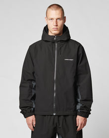 A short-haired person wears the LIVE FAST DIE YOUNG Basic Shell Jacket with reflektierender Druck and LIVE FAST on the chest, featuring an oversize fit, standing against a plain light gray background.