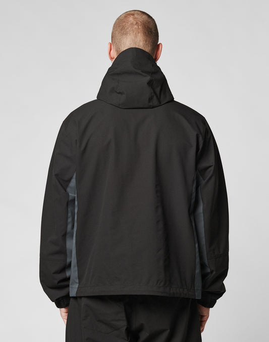 A person wearing the LIVE FAST DIE YOUNG Basic Shell Jacket, featuring an oversize fit, black color, dark gray side panels, and a water-repellent zipper, is shown from the back against a plain light background.