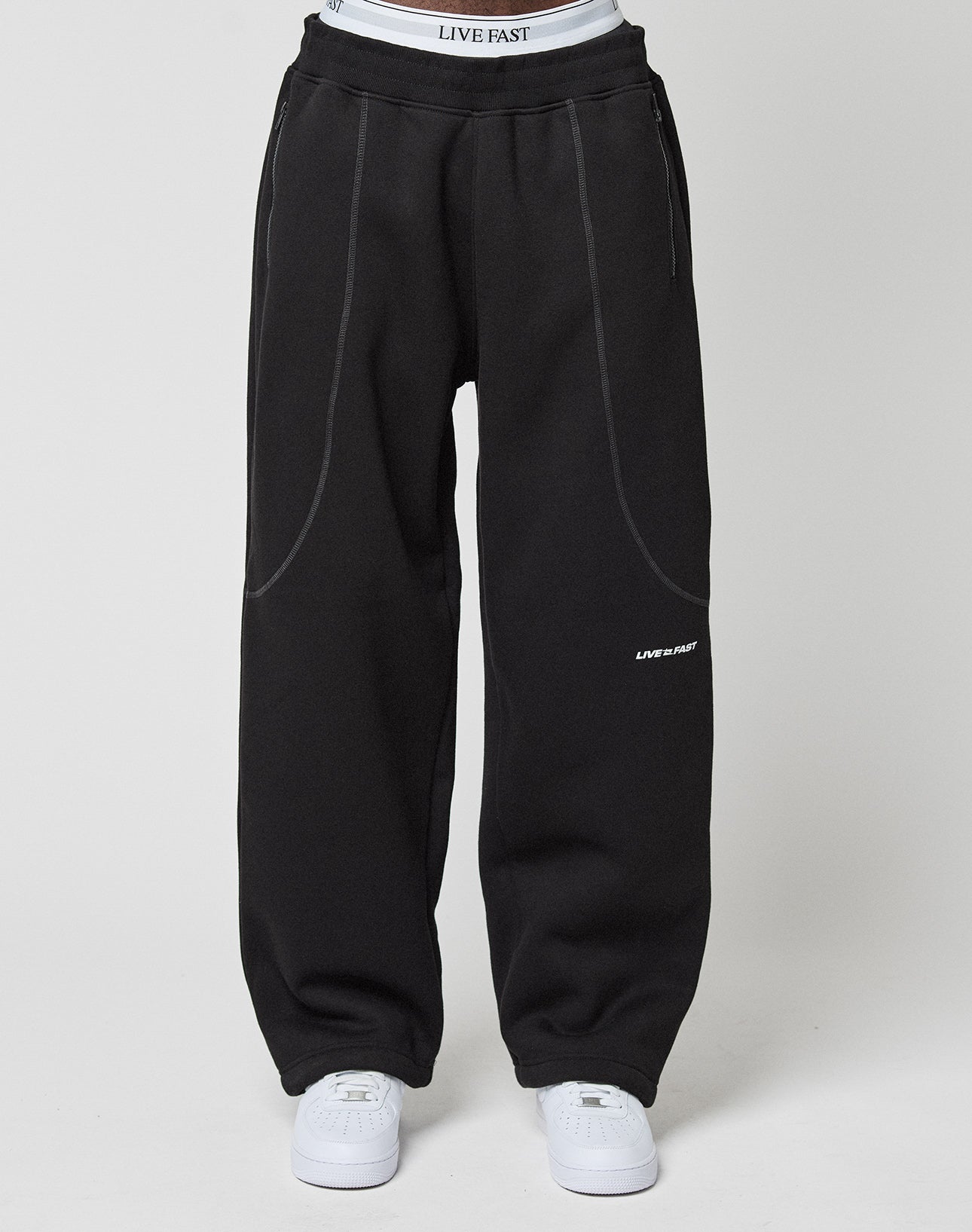 A person wears loose, wide-fit black LFDY Active Fleece Joggers by LIVE FAST DIE YOUNG with white stitching and a LIVE FAST logo on the thigh, paired with white sneakers and visible white LIVE FAST DIE YOUNG underwear above the waistband.