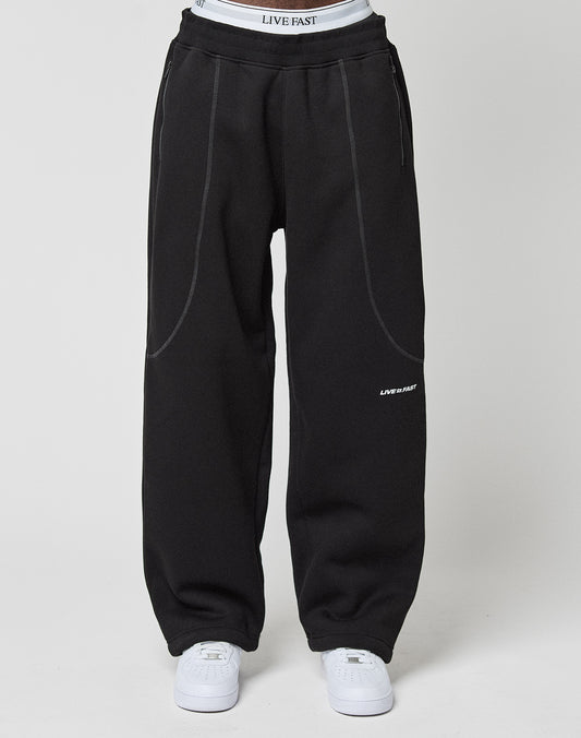 A person wears loose, wide-fit black LFDY Active Fleece Joggers by LIVE FAST DIE YOUNG with white stitching and a LIVE FAST logo on the thigh, paired with white sneakers and visible white LIVE FAST DIE YOUNG underwear above the waistband.