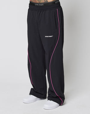 A person in LFDY Wavy Trackpants by LIVE FAST DIE YOUNG, wide fit black pants with pink piping and LIVE FAST logo on the elastic waistband and thigh, paired with white sneakers against a plain white background.