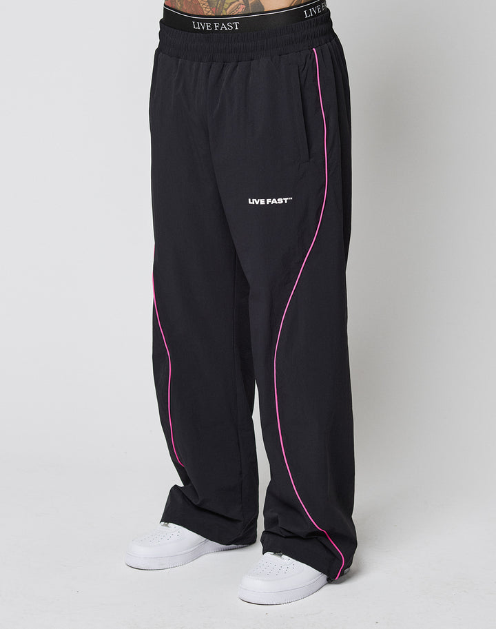 A person in LFDY Wavy Trackpants by LIVE FAST DIE YOUNG, wide fit black pants with pink piping and LIVE FAST logo on the elastic waistband and thigh, paired with white sneakers against a plain white background. | model
