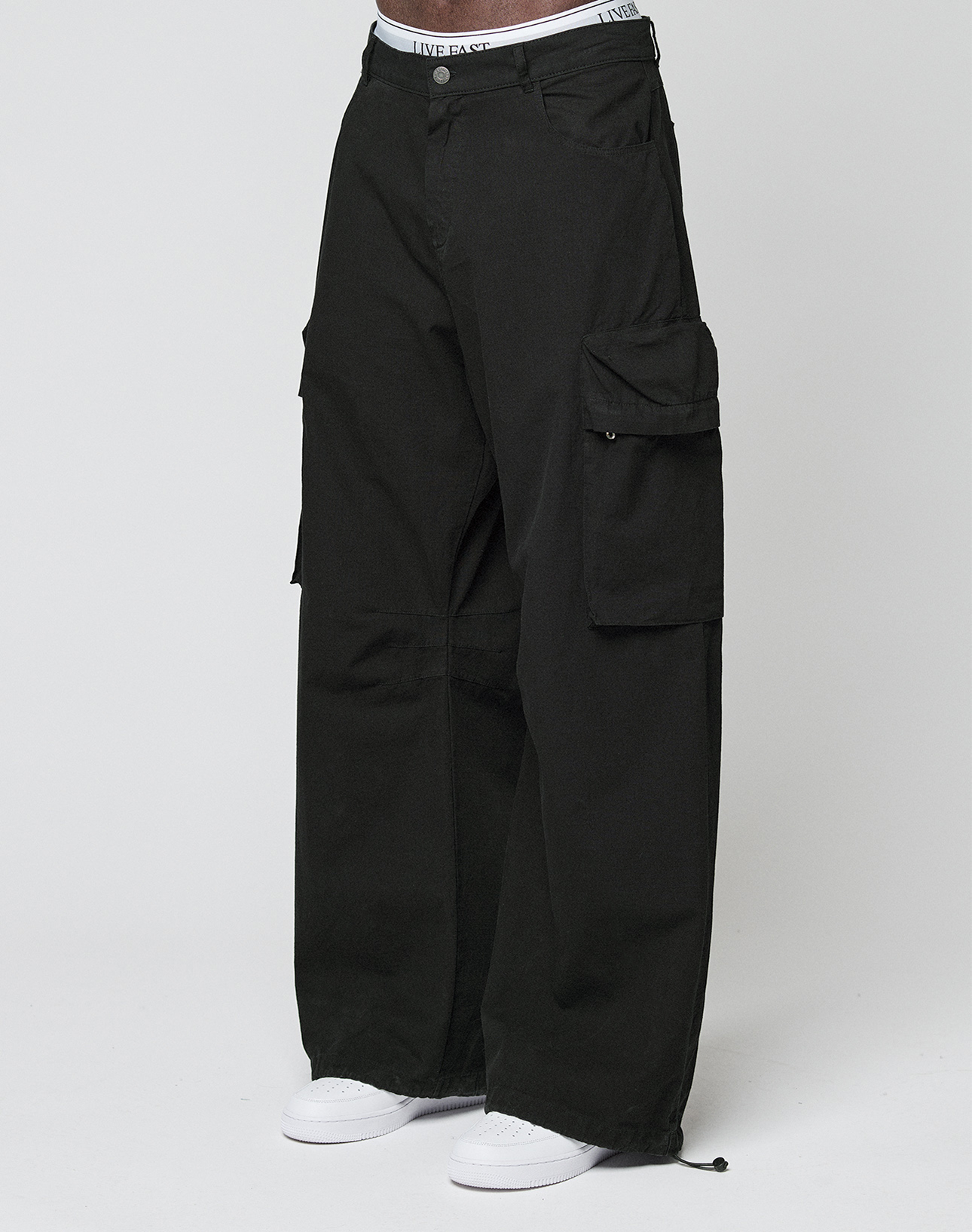 Eine Person trägt die LFDY Baggy Cargo von LIVE FAST DIE YOUNG aus 100% Baumwolle mit großen Cargo-Taschen, sichtbarem weißem Bund und weißen Sneakers vor einem schlichten, hellen Hintergrund.