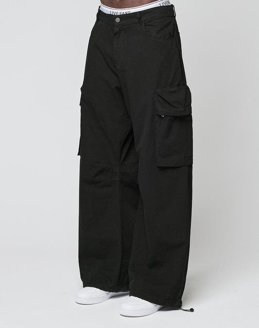 Eine Person trägt die LFDY Baggy Cargo von LIVE FAST DIE YOUNG aus 100% Baumwolle mit großen Cargo-Taschen, sichtbarem weißem Bund und weißen Sneakers vor einem schlichten, hellen Hintergrund.