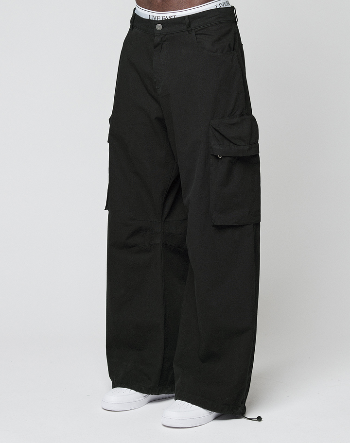 Eine Person trägt die LFDY Baggy Cargo von LIVE FAST DIE YOUNG aus 100% Baumwolle mit großen Cargo-Taschen, sichtbarem weißem Bund und weißen Sneakers vor einem schlichten, hellen Hintergrund.