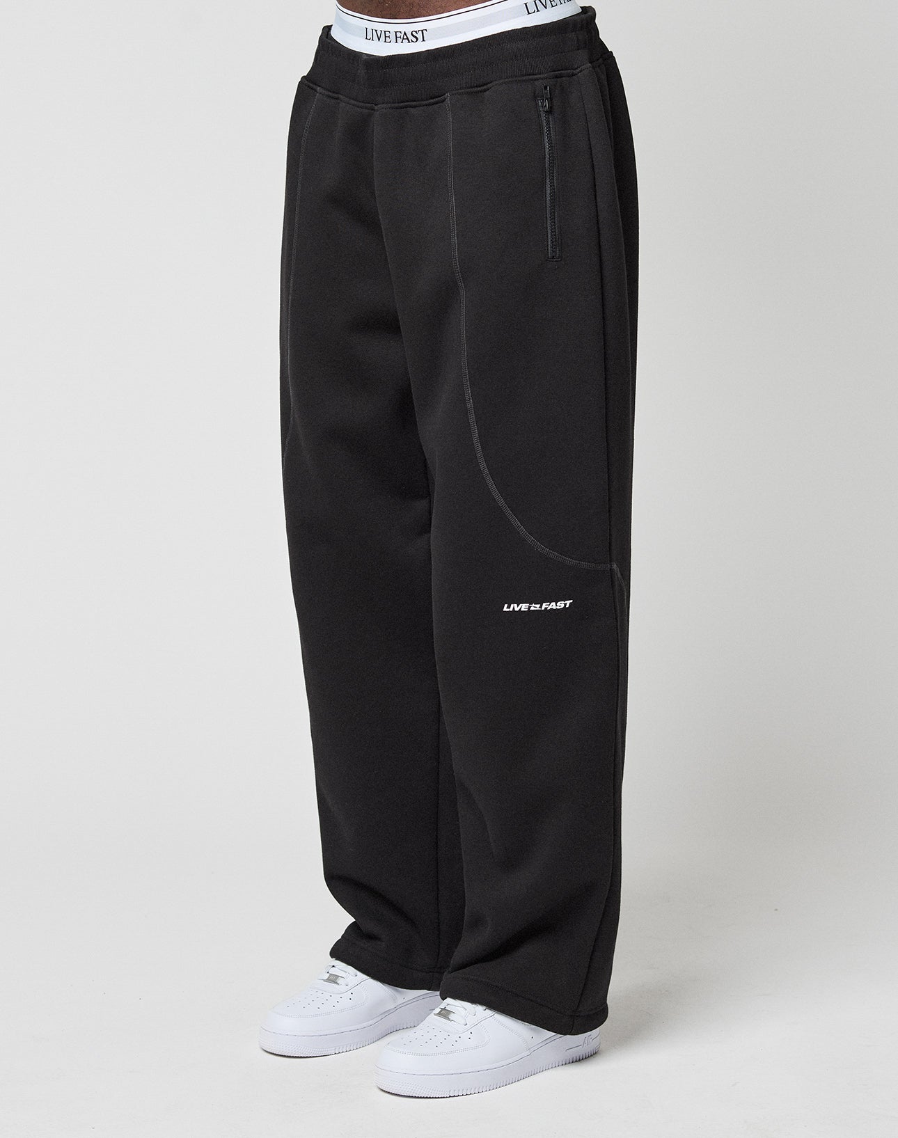 Model wears the LFDY Active Fleece Jogger by LIVE FAST DIE YOUNG—wide fit, black with LIVE FAST logo and branded elastic waistband—paired with white sneakers, standing against a plain white background.