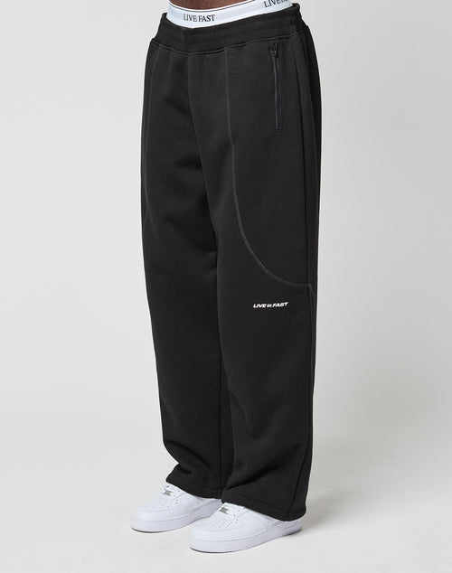 Model wears the LFDY Active Fleece Jogger by LIVE FAST DIE YOUNG—wide fit, black with LIVE FAST logo and branded elastic waistband—paired with white sneakers, standing against a plain white background.