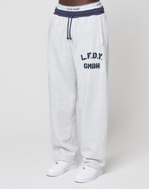 Wearing LFDY District Jogger by LIVE FAST DIE YOUNG: light gray wide-fit pants with “L.F.D.Y. GMBH” on the left thigh, dark gray contrast waistband with drawstrings, zip side pockets, white sneakers, and a “LIVE FAST” visible waistband.