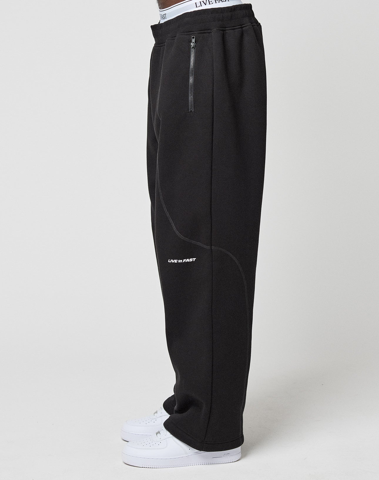 A person, shown from the waist down, models the LFDY Active Fleece Jogger by LIVE FAST DIE YOUNG—wide fit black sweatpants with zip pockets and a bold LIVE FAST logo on the thigh—paired with white sneakers against a plain background.