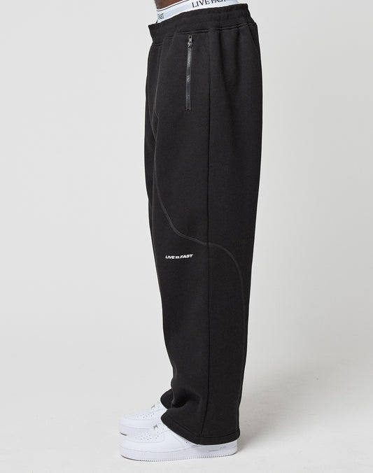 A person, shown from the waist down, models the LFDY Active Fleece Jogger by LIVE FAST DIE YOUNG—wide fit black sweatpants with zip pockets and a bold LIVE FAST logo on the thigh—paired with white sneakers against a plain background.