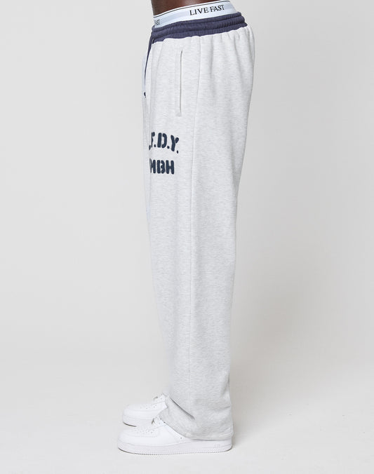 A person in profile wears LIVE FAST DIE YOUNG's LFDY District Jogger, light gray with wide fit, contrast waistband, “F.D.N.Y.” print on the side, and white sneakers, set against a plain light background.