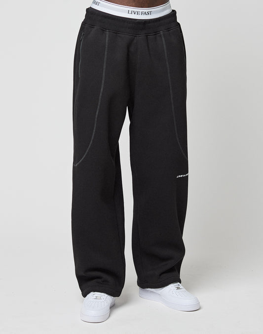 A person wears loose black LFDY Active Fleece Joggers by LIVE FAST DIE YOUNG, featuring subtle seam details and a Wide Fit. The white LIVE FAST waistband peeks above the pants, paired with white sneakers, against a plain light background.
