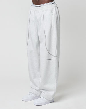 A person wears LFDY Active Fleece Jogger by LIVE FAST DIE YOUNG, featuring a wide fit, light gray color, curved seam details, zip pockets, logo print, and LIVE FAST text on the waistband and thigh, paired with white sneakers against a white background.