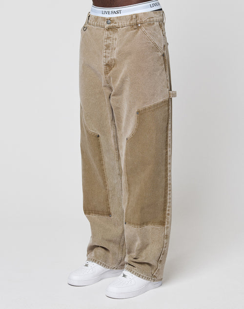 Someone stands against a plain light background wearing LIVE FAST DIE YOUNG Faded Carpenter Pants in a baggy fit, light brown color with visible seams, styled with white sneakers and a white LIVE FAST waistband.
