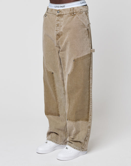 Someone stands against a plain light background wearing LIVE FAST DIE YOUNG Faded Carpenter Pants in a baggy fit, light brown color with visible seams, styled with white sneakers and a white LIVE FAST waistband.